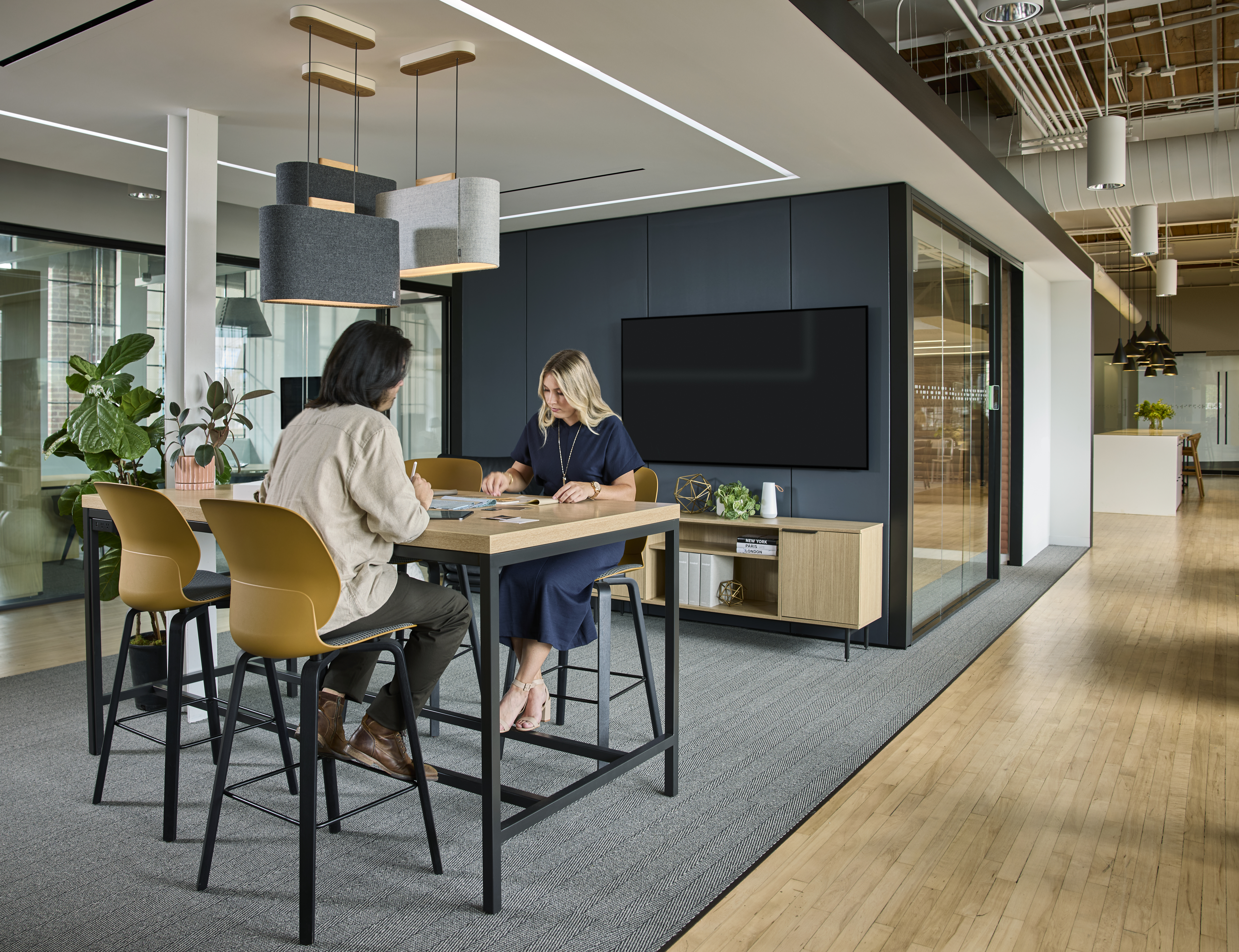 Two people meeting at a high-top table in a modern, open-concept office with warm finishes and acoustic lighting.